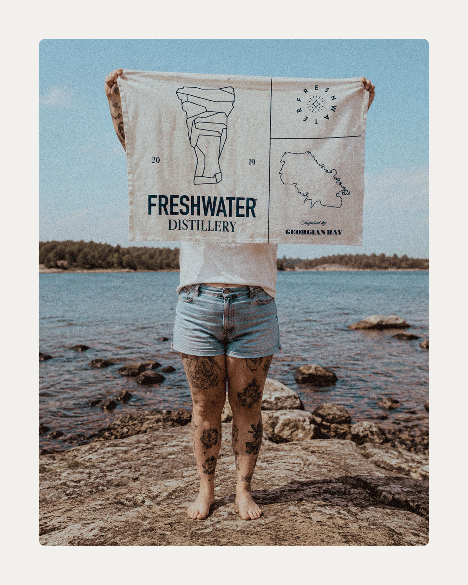 Person standing on the rocky Georgian Bay shoreline displaying a Freshwater Distillery flag.